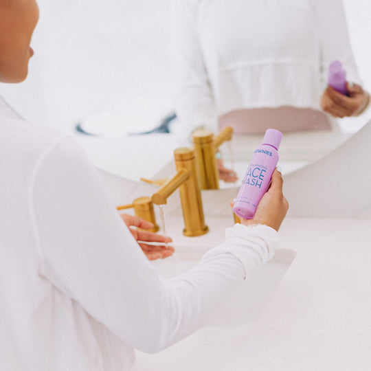 A woman in a bathroom holding Frownies face wash, standing in front of a mirror with half of her reflection visible.