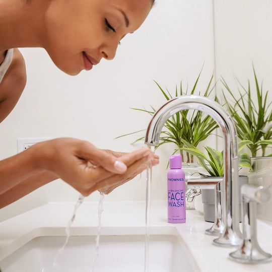 A woman leaning over a bathroom counter with her hands under a running sink, catching and holding water as if about to wash her face, with Frownies face wash visible in the background.