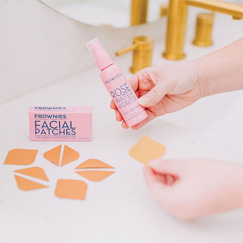 Facial patches and rose water shown being held by hands by a bathroom sink. The Frownies box and patches are also shown laying on the counter