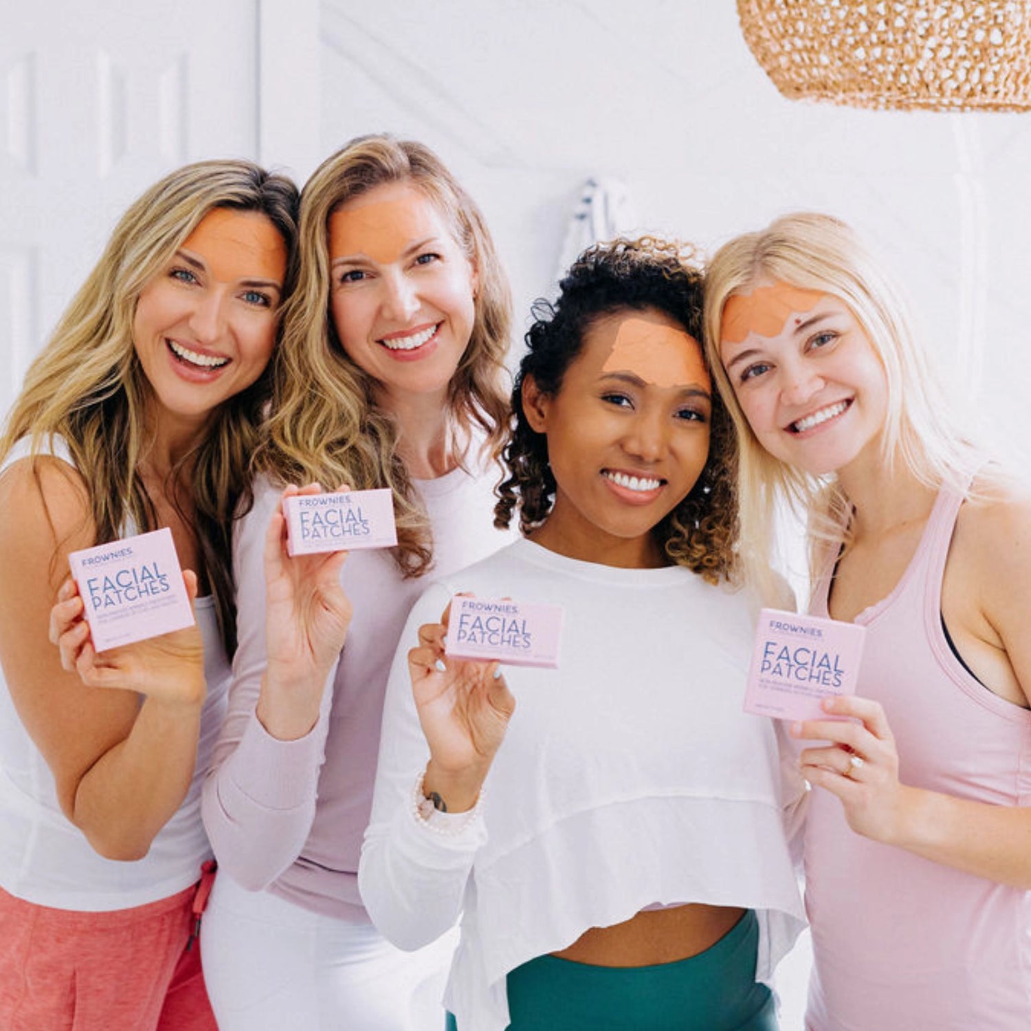 Four women smiling while holding Frownies facial patches and wearing the facial patches on their faces.