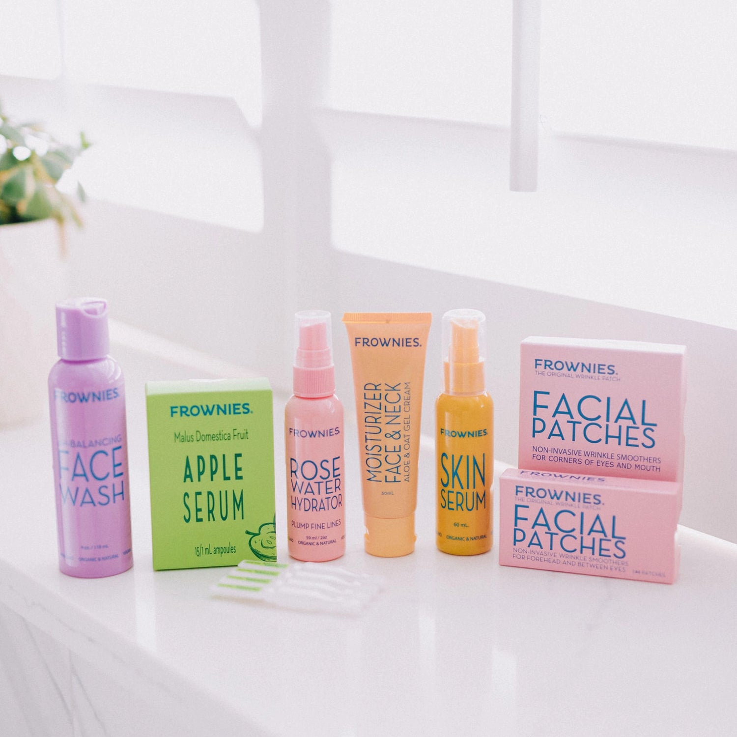 Frownies products displayed together, including face wash, apple serum, rose water hydrator, moisturizer, skin serum, and facial patches (FBE and CEM)