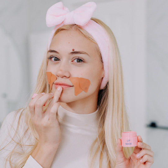 A woman shown putting on frownies lip mask while also wearing frownies facial patches and skincare headband. She is holding the lip mask container in her hand as well