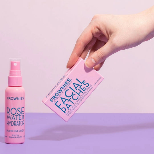 Frownies Forehead and Between Eyes (FBE) facial patches held by a hand, shown almost being set down next to a Frownies rose water hydrator bottle, with a lilac and dual pinkish color background.