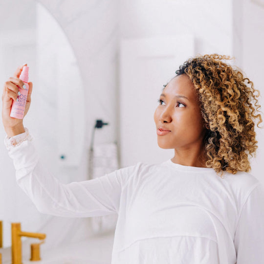 A model with curly hair spraying rose water hydrator onto her face.