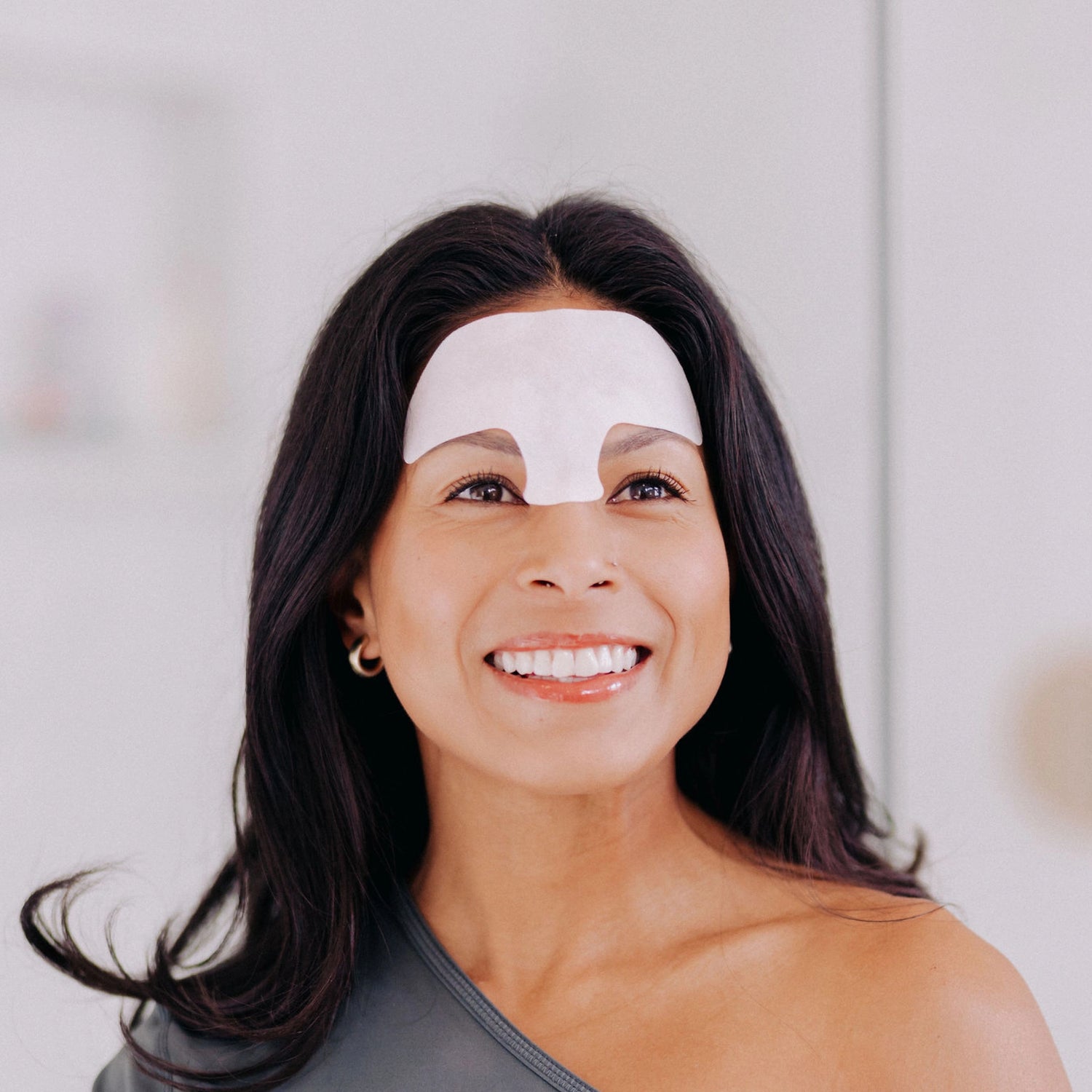 A woman with black hair smiling while wearing a Frownies forehead serum patch.