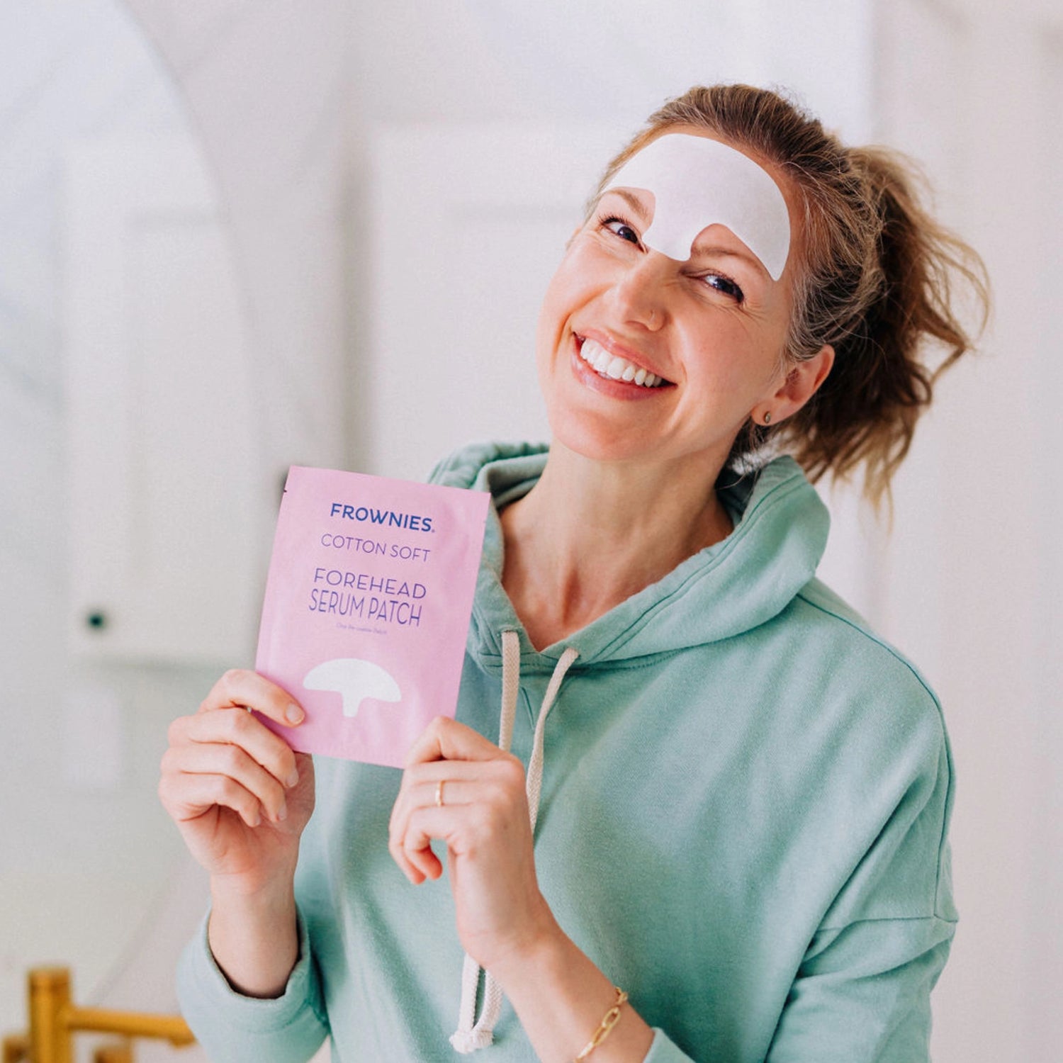 A smiling woman wearing a Frownies forehead serum patch, holding up the packaging for the camera to see.