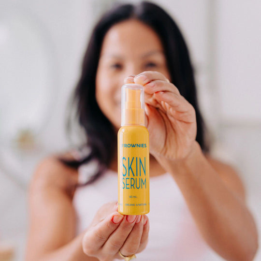 Close-up of a smiling woman with a blurred face, holding up a Frownies skin serum bottle in focus in front of her face.