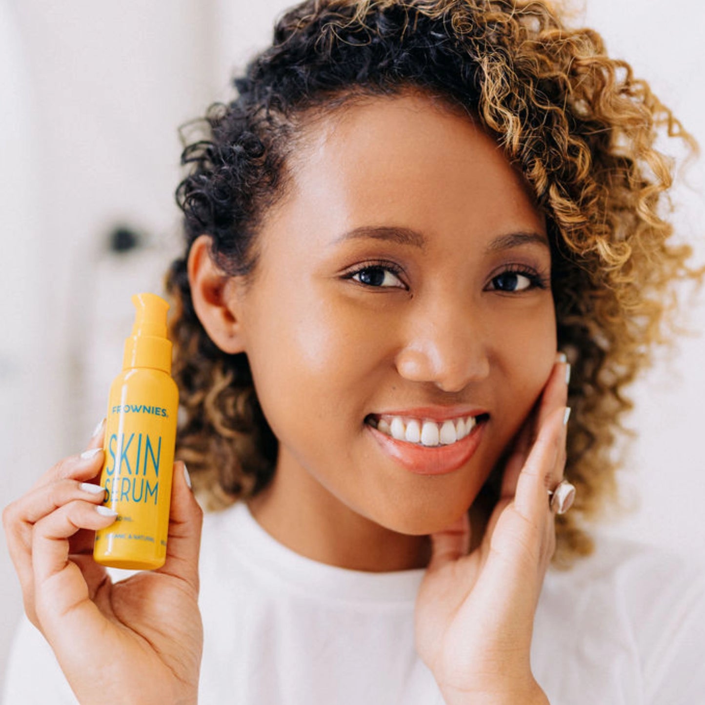 A curly-haired Black woman smiling, gracefully touching her face with her left hand while holding Frownies moisturizer for face and neck in her right hand.