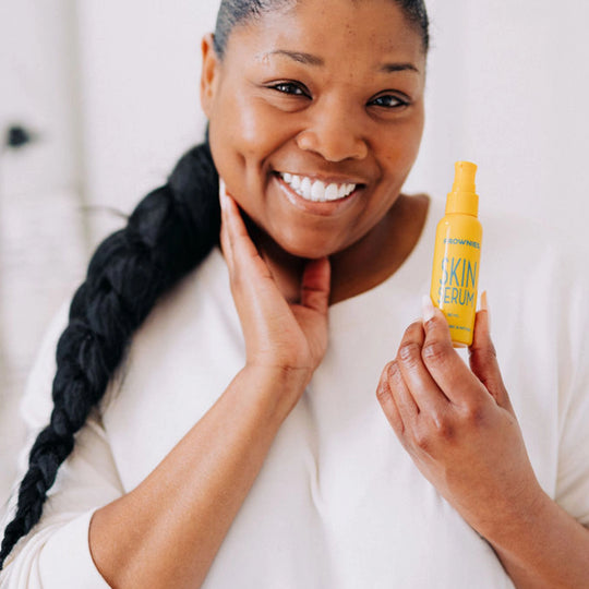 Close-up of a woman smiling with one hand behind her head, holding Frownies skin serum in her other hand, clearly displaying the product packaging.