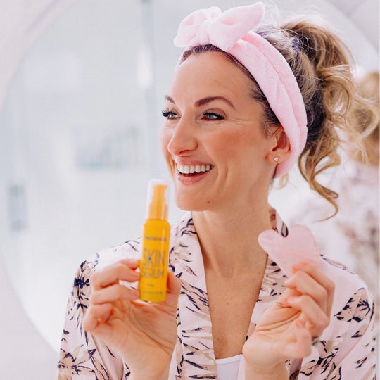 Woman smiling and looking to her right, wearing a Frownies facial care headband and holding a Frownies Skin Serum bottle and Gua Sha stone, clearly displaying the products.