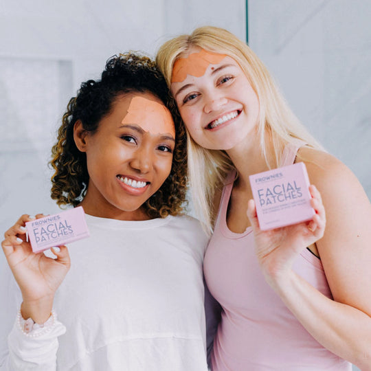 Two women smiling while wearing facial patches on their forehead, holding the product packaging visibly in their hands.