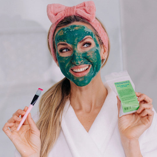 Happy woman shown in the mirror with Frownies Blue Green Algae Mask applied to her face, holding the product packaging and brush in her hands while wearing a Frownies skincare headband.