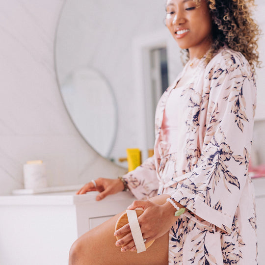 Curly-haired woman in a robe using a Frownies body brush on her leg.